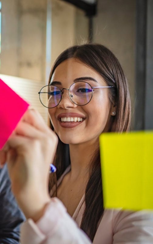 A woman with long brown hair and glasses smiles while writing on a bright pink sticky note at a LATAM Talent Hub